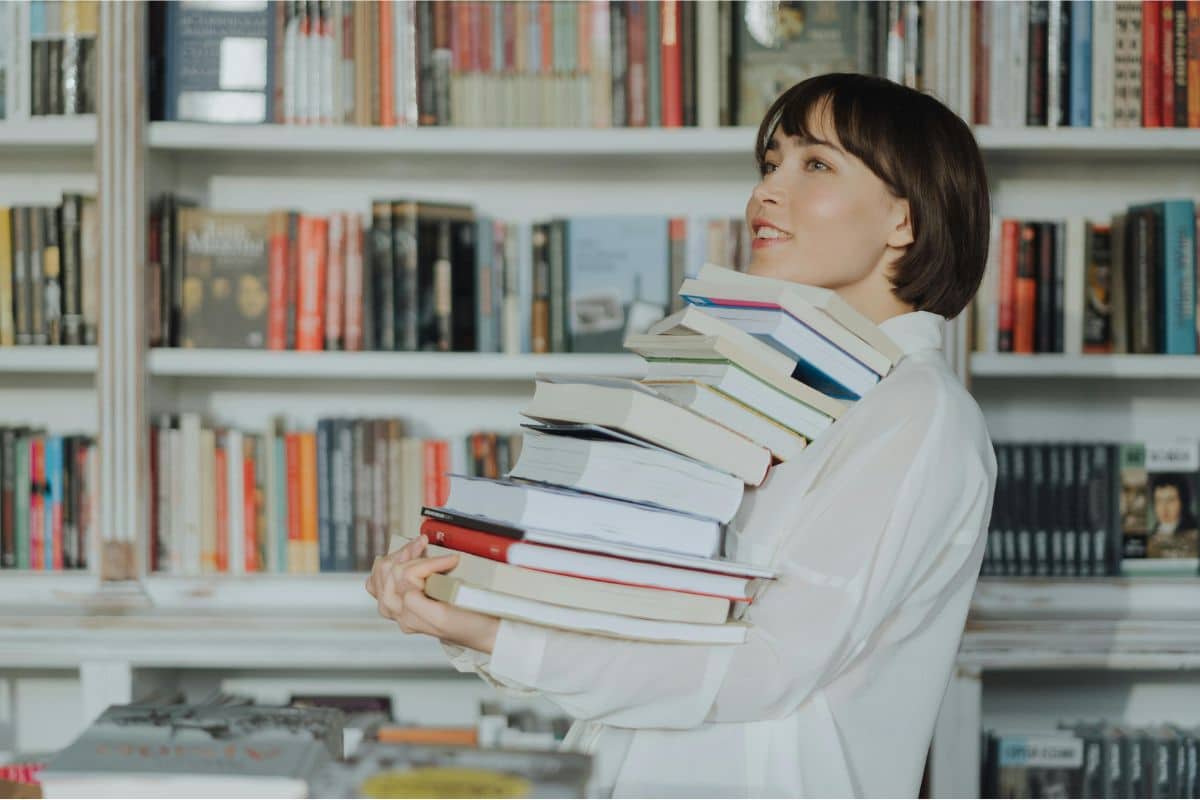 Frau mit einem Stapel Büchern in der Armen vor einem Bücherregal